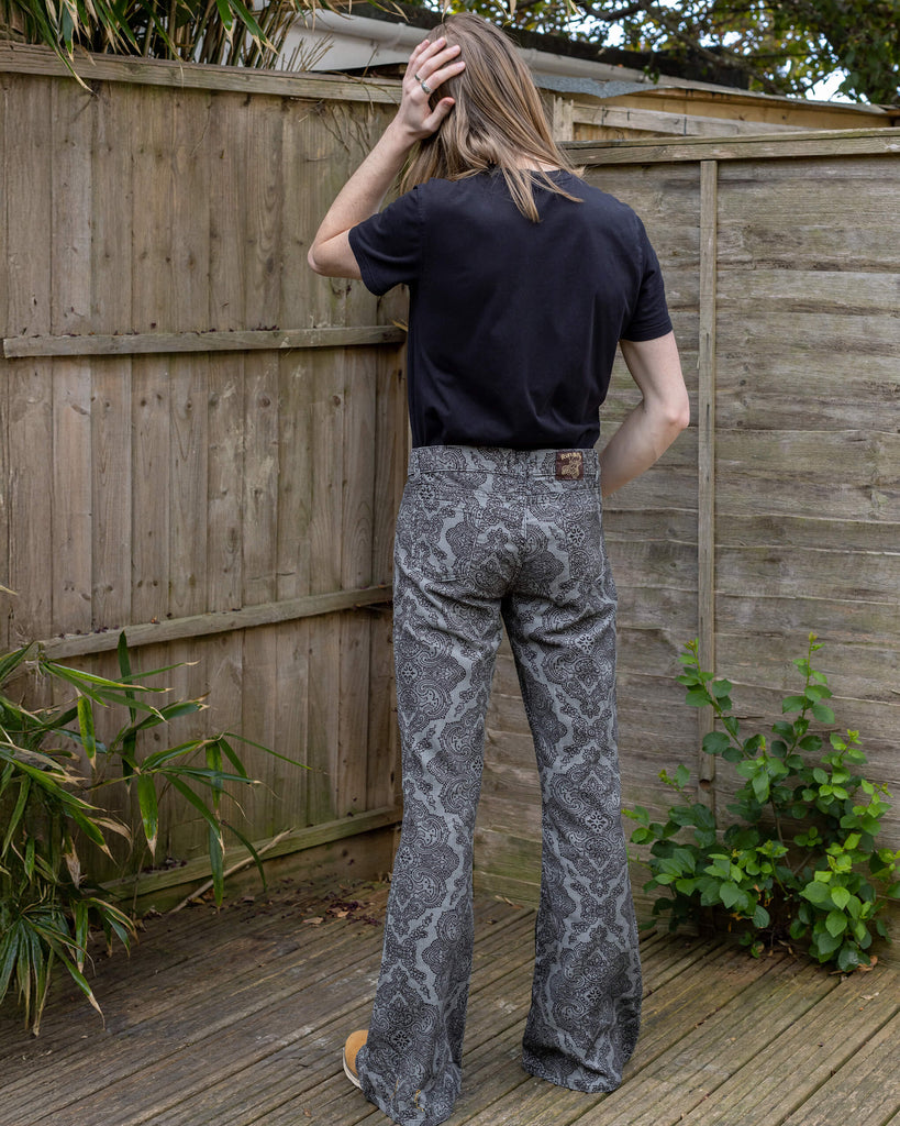 Model Jack is wearing Run & Fly’s Silver Hendrix flares, shown from the back in an outdoor garden setting with a wooden fence. The flares have a detailed black paisley pattern on a silver background.
