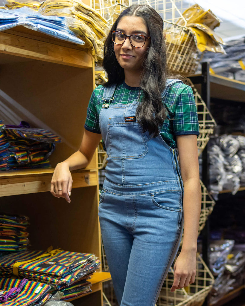 Model is stood in our warehouse wearing the light blue denim stretch skinny flared dungarees with a green tartan tshirt and black trainers. They are posing leaning on the shelves with one leg crossed over the other and smiling.
