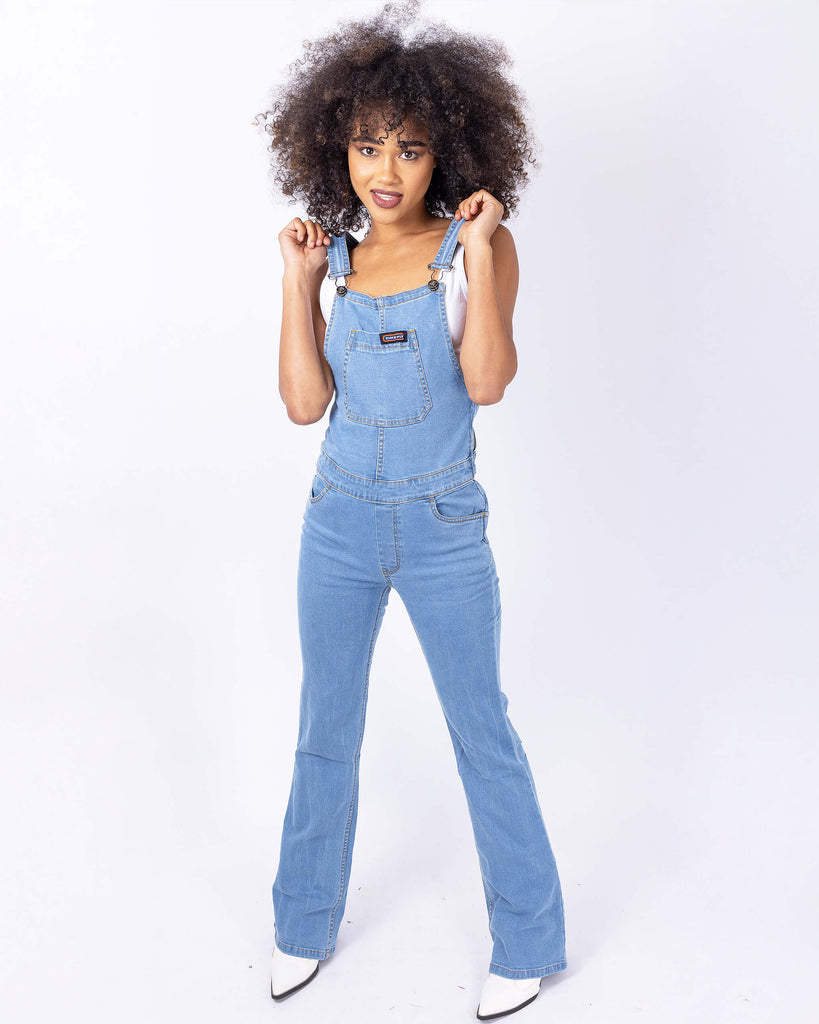 Cindy is stood in front of a white studio background wearing the light blue denim stretch skinny flared dungarees with a white short sleeved tshirt underneath and white cowboy boots. They are facing the camera with both hands up holding onto the shoulder straps.