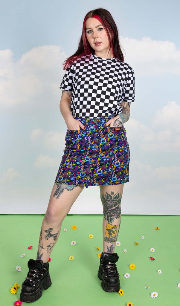Florence is stood in a photography studio in Hove in front of blue sky backdrop on a green floor with flowers scattered wearing 90's Arcade A Line Skirt with black and white checker board short sleeve tee and black shoes. The skirt print features classic 90's style shapes, squiggles and doodles in green, orange, pink, purple and light blue all on a dark blue background. Florence is posing with her hands in the skirt pockets.