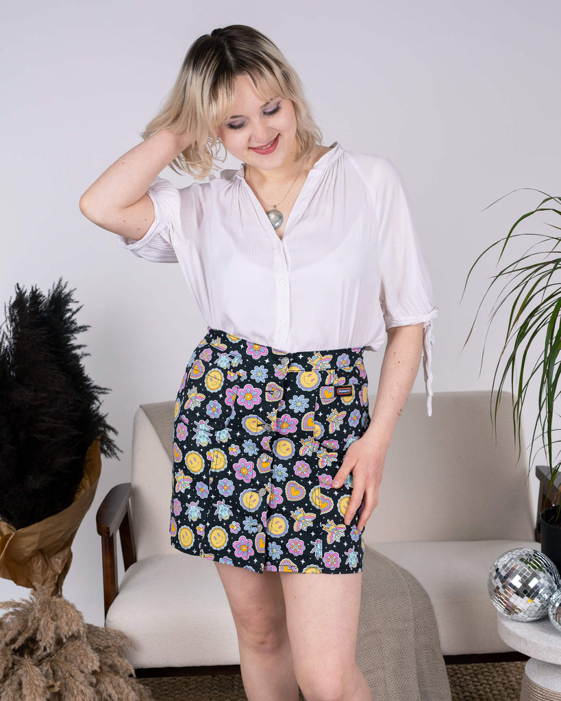 Model adjusts their hair while showcasing the Bee Happy A-Line Skirt by Run & Fly. The skirt features a cheerful black design with smiley suns, vibrant flowers, bees, and hearts. Styled with a breezy white blouse, highlighting the skirt’s colourful personality. Ethical slow fashion made with care.