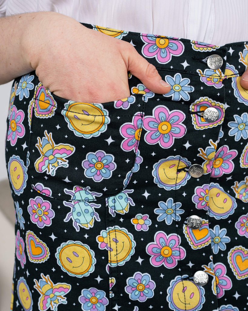 Detailed close-up of the Bee Happy A-Line Skirt by Run & Fly, highlighting the cheerful print of colourful bees, flowers, hearts, and suns. The silver buttons and pocket detail stand out. Paired with a white blouse for a fresh look. Made by a small ethical slow fashion brand.