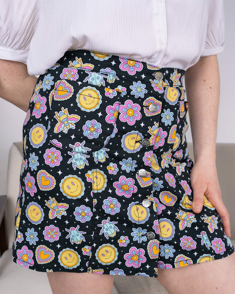Close-up view of the Run & Fly Bee Happy A-Line Skirt, showcasing its vibrant print featuring smiley suns, flowers, hearts, and cute bees on a black background. The skirt features visible silver buttons and practical pockets. Model wears a white blouse, focusing on the skirt’s intricate details. Ethical slow fashion by Run & Fly.