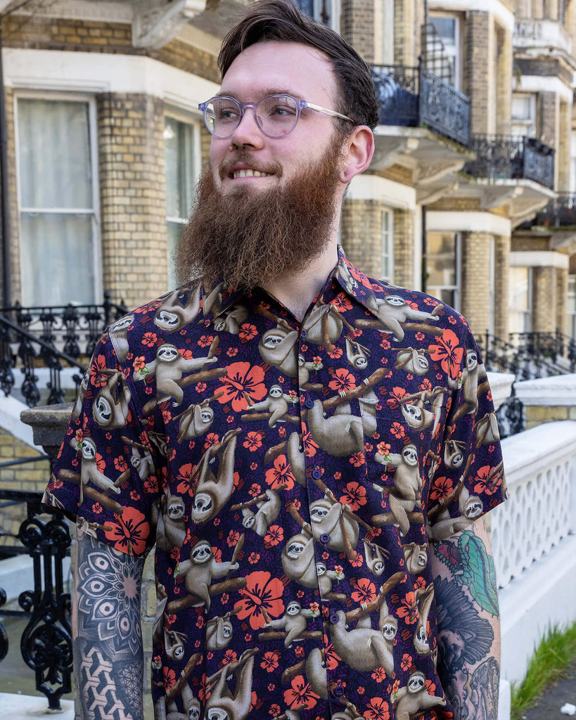 Declan a male in his 20's with a bears and glasses is wearing a sloth shirt with Hawaiian flowers on and beige cargo trousers on a street in Hove.