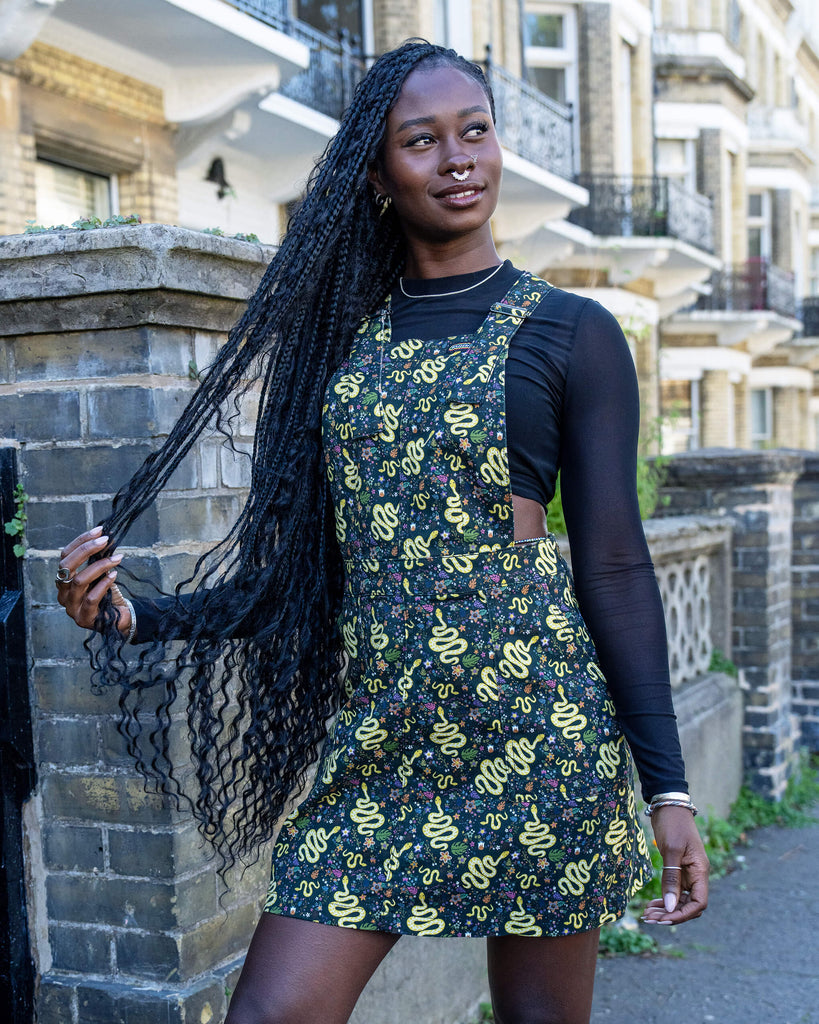 A model is stood outside in Hove wearing Run & Fly Halloween: Serpent Garden Stretch Twill Pinafore Dress with a long sleeved black mesh top underneath. The dark base of the pinafore is covered with vibrant yellow snake designs, surrounded by small floral patterns, creating a quirky, eye-catching look. The model pairs the outfit with black high-top trainers and is smiling at the camera with her hands in her hair