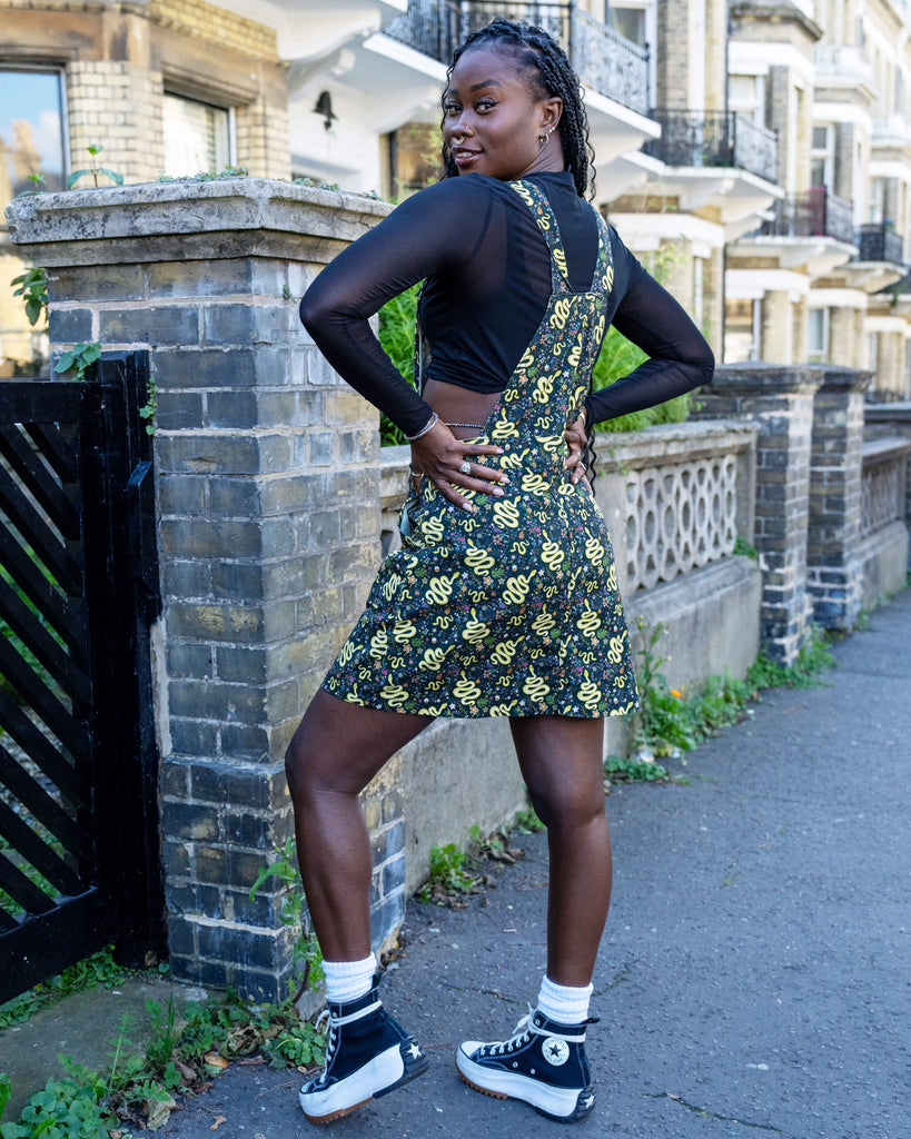 A model is stood outside in Hove wearing Run & Fly Halloween: Serpent Garden Stretch Twill Pinafore Dress with a long sleeved black mesh top underneath. The dark base of the pinafore is covered with vibrant yellow snake designs, surrounded by small floral patterns, creating a quirky, eye-catching look. The model pairs the outfit with black high-top trainers and is stood with their back to the camera with one leg slightly bent and is looking back over her shoulder at the camera 