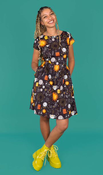 Milly, a femme model with locs, is wearing a black belted tea dress with an all over print of Katie Abey witchy illustrations with a matching fabric belt tied across the waist. The dress has been paired with bright green boots. Milly is smiling at the camera and the background of the photo is a forrest green. 