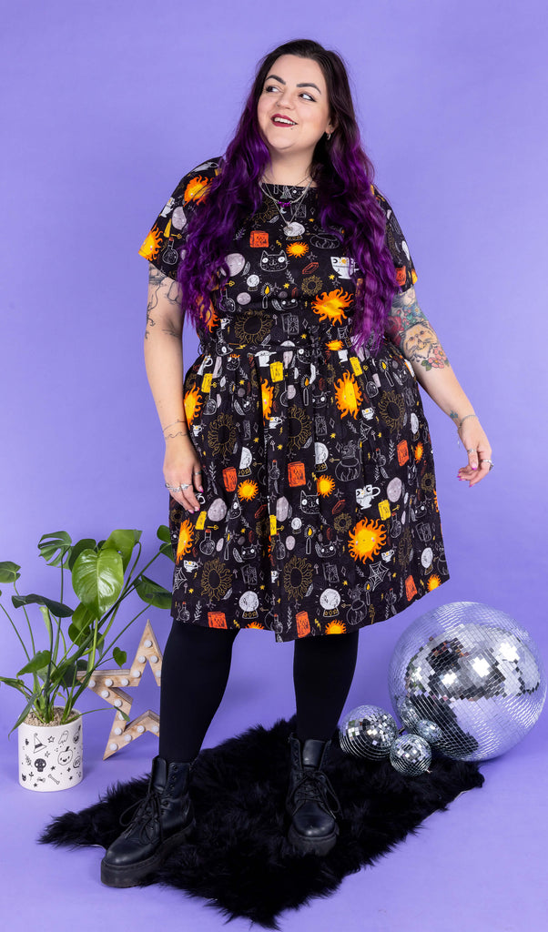 Luisa, a femme model with purple hair and tattoos, is stood on a furry black rug amongst disco balls, a star light and a plant in a photography studio in Hove wearing Run & Fly x Katie Abey Solar Witch Stretch Belted Tea Dress with Pockets with black tights and boots. The black dress has an all over witchy print illustrated by Katie Abey. Luisa is posing in front of a lilac backdrop with her arms by her sides and one leg in front of the other and smiling looking off to one side.