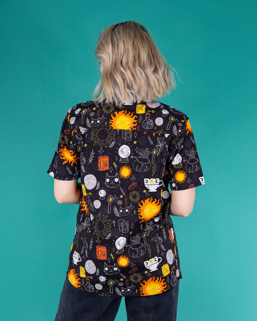 Amy a femme white bobbed hair looking into camera wearing the Katie Abey Solar Witch Shirt shot in Amy Davies Photography studio in Hove she is facing away to show the back. The print is a black base with Katie abey witchy style illustrations of suns, potions, books, cats, sparkles and more.
