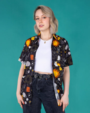 Amy a femme white bobbed hair looking into camera wearing the Katie Abey Solar Witch Shirt shot in Amy Davies Photography studio in Hove. The print is a black base with Katie abey witchy style illustrations of suns, potions, books, cats, sparkles and more.