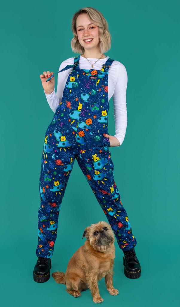 Amy a blonde white femme model with shoulder length hair is wearing a white long sleeved top with black boots and dogs in space blue dungarees featuring dogs in spaceships, planets, and stars on a blue base. She is joined by Brian a Brussels Griffon