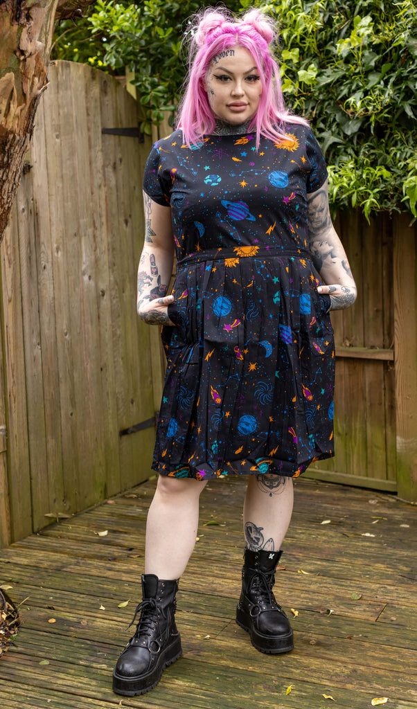 Zoe is a tattooed white female with pink long hair and is wearing a galaxy print space planets tea dress in a garden setting