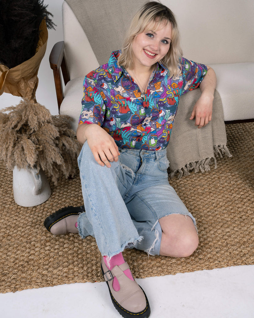 Run & Fly Spooky Stuff Short Sleeve Shirt modelled with light blue distressed denim jeans. The colourful spooky-themed print includes ghosts, cauldrons, and creepy-cute details, bringing a fun and quirky flair. The model is seated, exuding relaxed confidence and a cheerful smile. Run & Fly’s ethical slow fashion designs are perfect for expressing individuality with a Halloween twist.