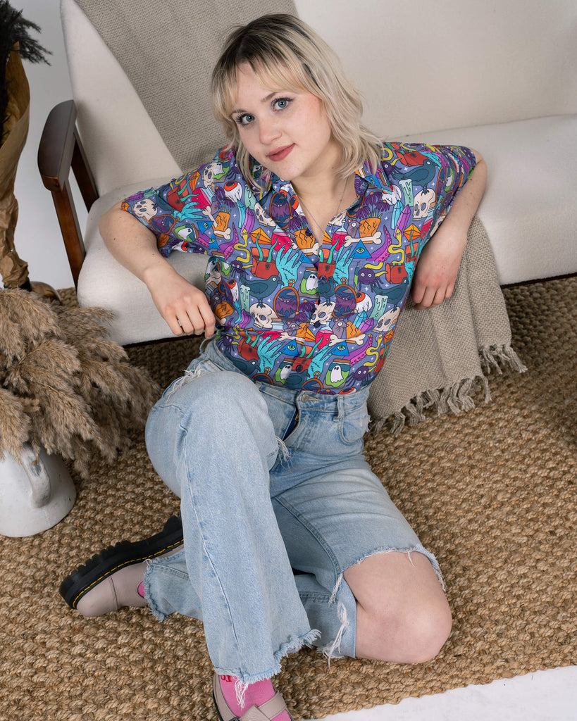 Model showcasing the Run & Fly Spooky Stuff Short Sleeve Shirt with a vibrant Halloween-themed design. The shirt features ghosts, skulls, and witchy motifs in bold colours, styled with distressed light blue jeans. Posed sitting on a cosy rug, the model’s outfit captures a relaxed, playful aesthetic. Perfect for embracing spooky season or adding a quirky touch to your everyday wardrobe.