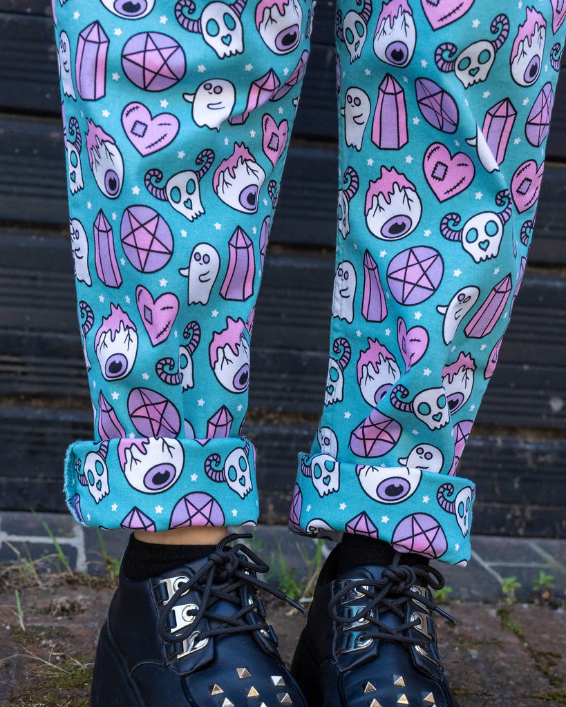 A close-up shot of the legs and feet, highlighting the turquoise dungarees with a pastel pink and purple spooky print. The woman wears black platform shoes with silver spikes on the front, adding an edgy touch to the whimsical print of the dungarees by Run & Fly.