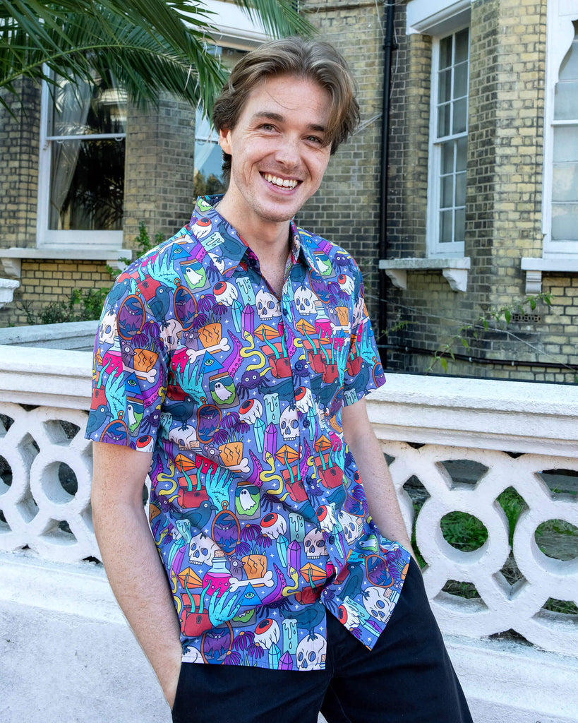 Jack is stood outside in Hove wearing Run & Fly Halloween: Spooky Stuff Short Sleeve Shirt with dark trousers. The purple button up shirt has an all over print of various Halloween themed items such as skulls, snakes, eyeballs, ghosts, bats, crows, candles, potions, crystals and more. He is smiling at the camera with his hands in his pockets. 