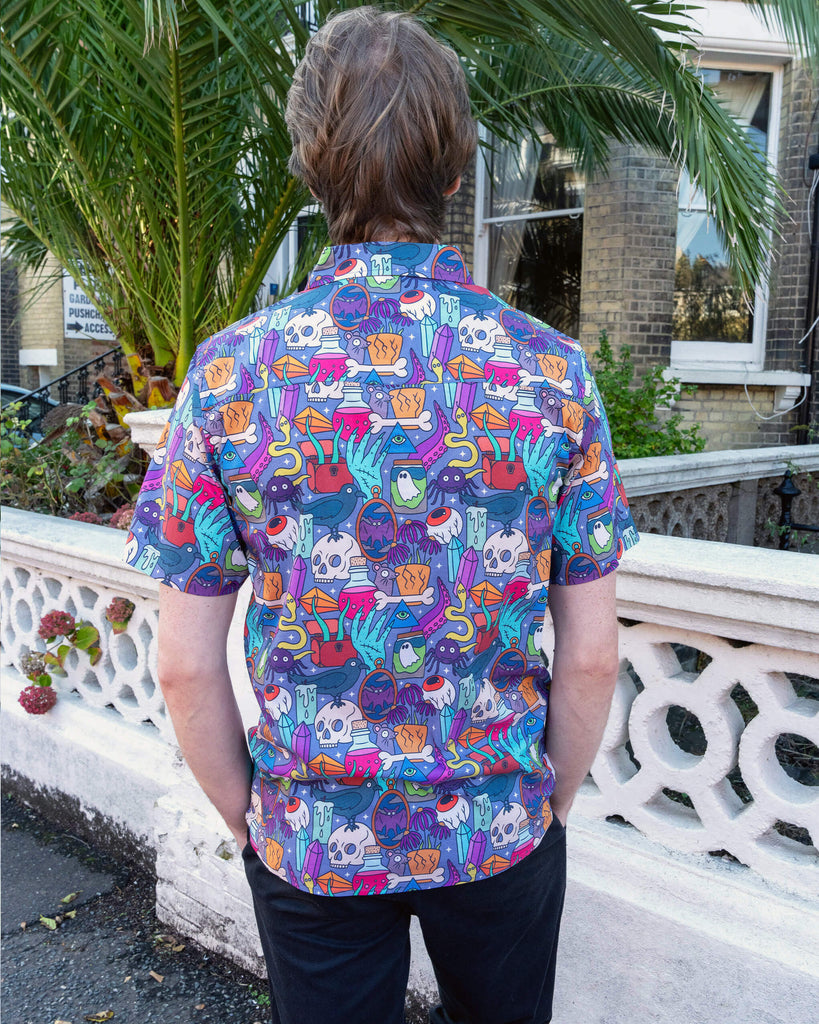 Jack is stood outside in Hove wearing Run & Fly Halloween: Spooky Stuff Short Sleeve Shirt with dark trousers. The purple button up shirt has an all over print of various Halloween themed items such as skulls, snakes, eyeballs, ghosts, bats, crows, candles, potions, crystals and more. Jack is stood with his back to the camera to show the back of the shirt with his hands in is pockets.