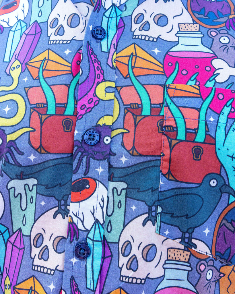 Close up of model wearing Run & Fly Halloween: Spooky Stuff Short Sleeve Shirt with dark trousers. The purple button up shirt has an all over print of various Halloween themed items such as skulls, snakes, eyeballs, ghosts, bats, crows, candles, potions, crystals and more.