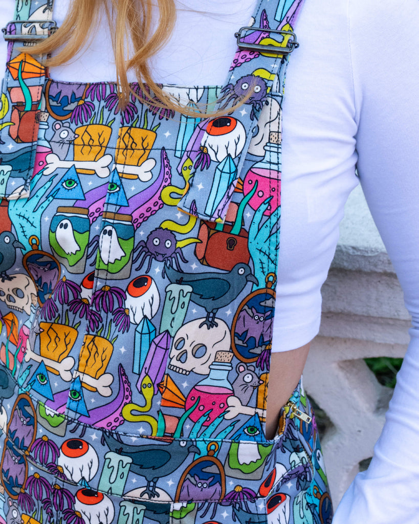 Close up of Katie stood outside in Hove wearing Halloween: Spooky Stuff Stretch Twill Pinafore Dress with a long sleeved cropped white top underneath. The light blue pinafore has an all over print of various Halloween themed items such as skulls, snakes, eyeballs, ghosts, bats, crows, candles, potions, crystals and more. Katie is posing toward the camera with he hair swishing in the wind.