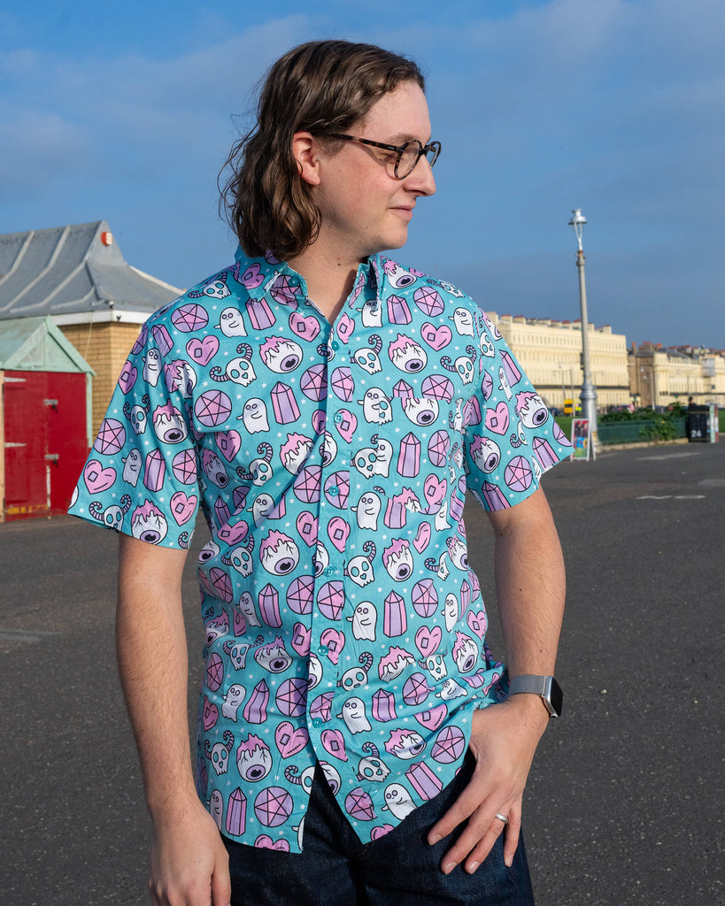 Model posing in Run & Fly's ethically made Spooky Stuff shirt. The unique print includes pastel pink ghosts, crystal motifs, and stitched hearts on a teal background, ideal for fans of quirky alternative style.