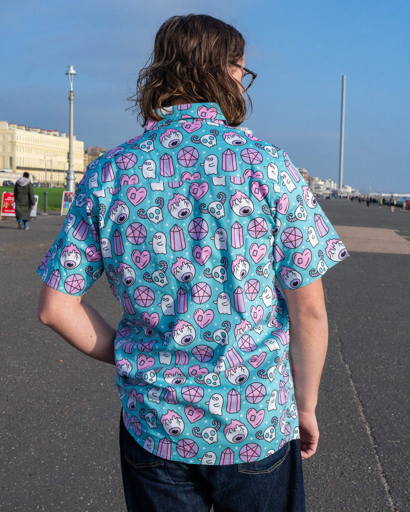 Back view of a model wearing Run & Fly's Spooky Stuff short-sleeved shirt. The ethically made design features pastel pink and blue ghosts, skulls, pentagrams, and crystals on a vibrant teal background, perfect for alternative fashion lovers.
