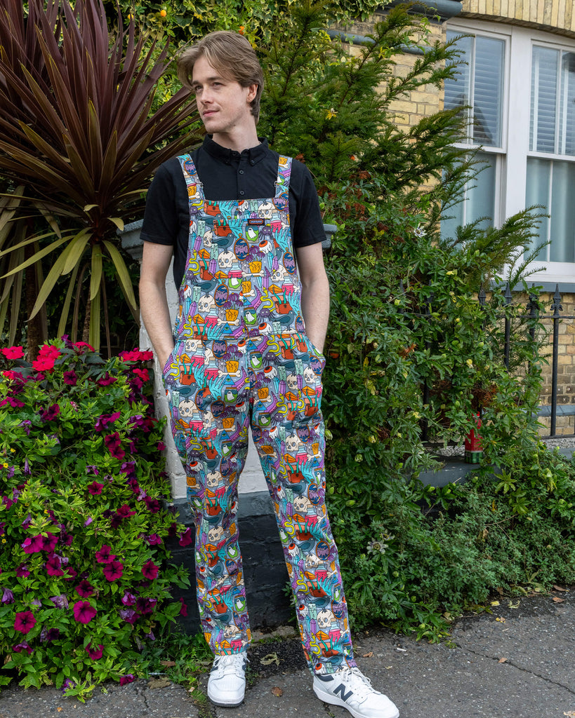 Jack is stood outside in Hove by greenery  wearing Run & Fly Halloween: Spooky Stuff Stretch Twill Dungarees with a short sleeve black top underneath. The light blue dungarees have an all over print of various Halloween themed items such as skulls, snakes, eyeballs, ghosts, bats, crows, candles, potions, crystals and more. Jack is smiling facing the camera with his hands in his pockets. 