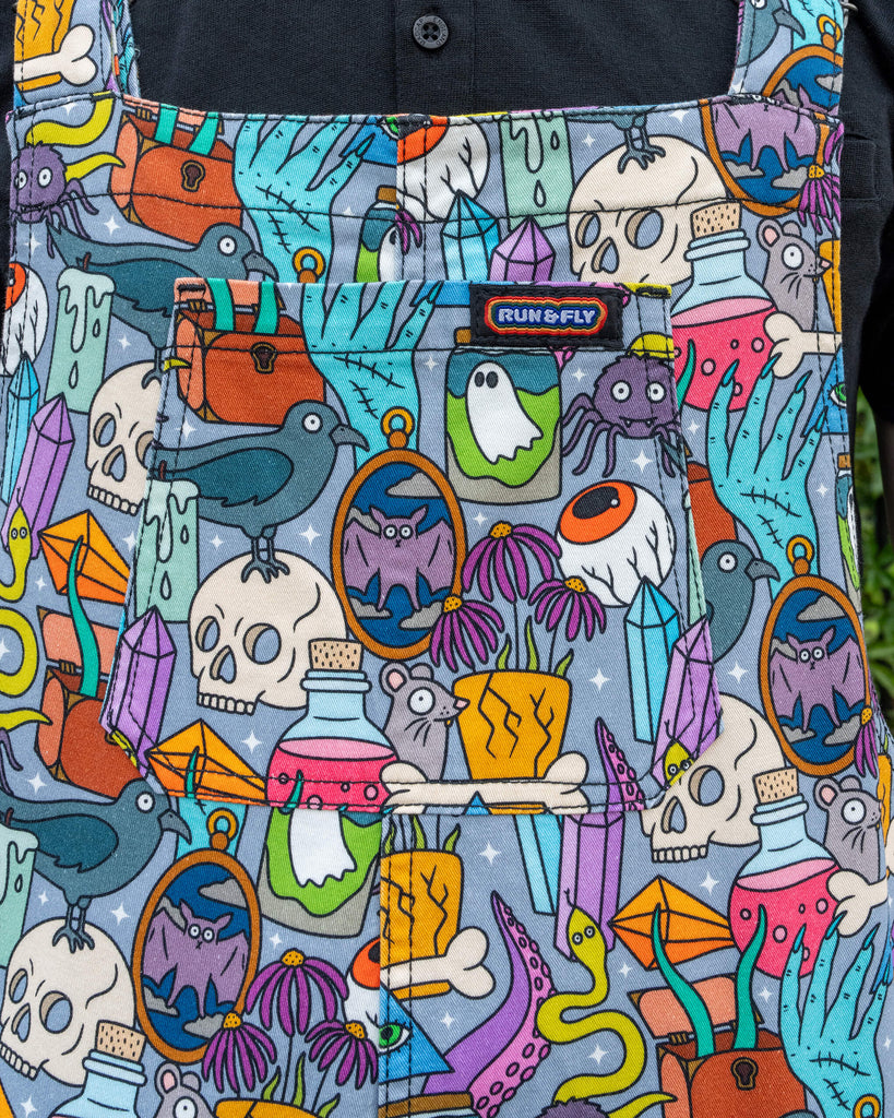 Close-up of Run & Fly’s “Spooky Stuff” dungarees, featuring a colourful Halloween-inspired print designed by Waffle Mama. The design includes playful elements such as skulls, ravens, ghosts, potions, and crystals, all set against a grey background. The detailed pattern highlights the brand’s fun and quirky approach to ethical slow fashion.
