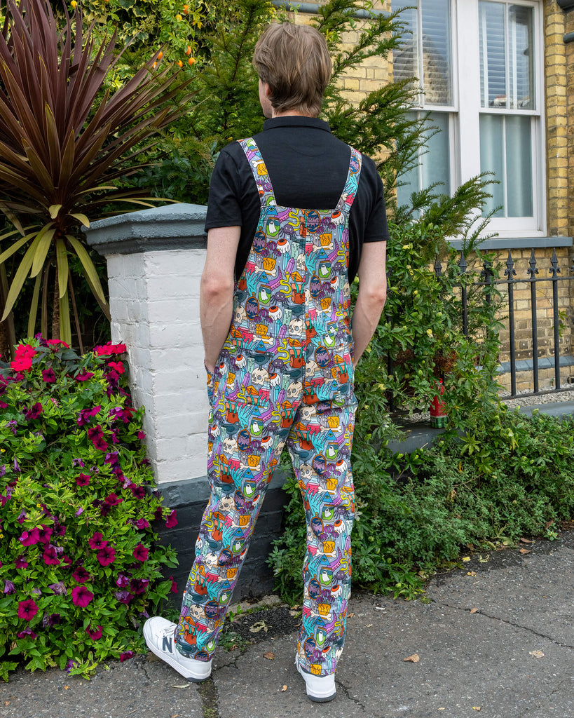Jack is stood outside in Hove by greenery  wearing Run & Fly Halloween: Spooky Stuff Stretch Twill Dungarees with a short sleeve black top underneath. The light blue dungarees have an all over print of various Halloween themed items such as skulls, snakes, eyeballs, ghosts, bats, crows, candles, potions, crystals and more. He is stood with his back to the camera to show the back of the dungarees and his hands are in his pockets.