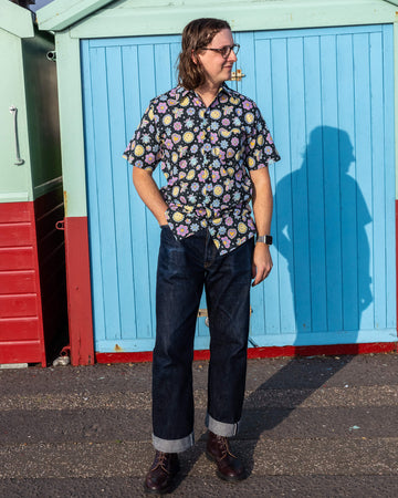 Michael is stood outside in front of a blue beach hut in Hove wearing Run & Fly Bee Happy short sleeve shirt with dark blue jeans and brown shoes. The black shirt has an all over colourful bee, smiley face and flower print. They are posing look off to one side with their hand in their pocket. 