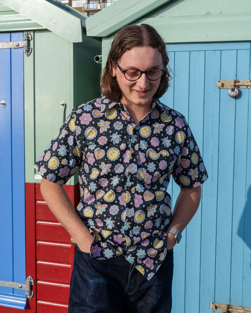 Michael is stood outside in front of a blue beach hut in Hove wearing Run & Fly Bee Happy short sleeve shirt with dark blue jeans. The black shirt has an all over colourful bee, smiley face and flower print. Michael is looking down smiling with his hands in his pockets.