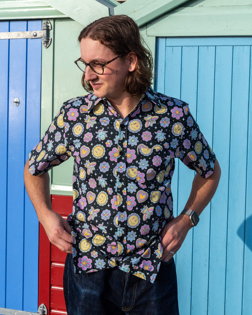 Michael is stood outside in front of a blue beach hut in Hove wearing Run & Fly Bee Happy short sleeve shirt with dark blue jeans. The black shirt has an all over colourful bee, smiley face and flower print. Michael is posing with his hands on his hips and looking off to one side.