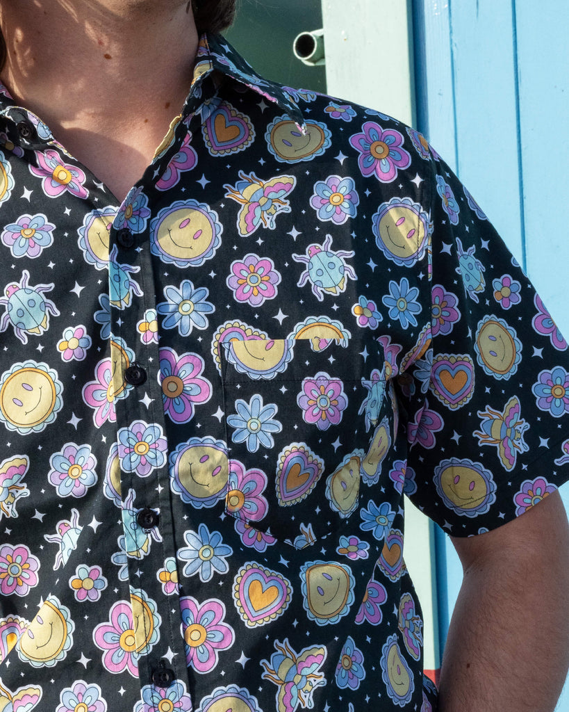 Close up of model wearing Run & Fly Bee Happy short sleeve shirt with dark blue jeans. The black shirt has an all over colourful bee, smiley face and flower print. Close up shows the front pocket on the shirt