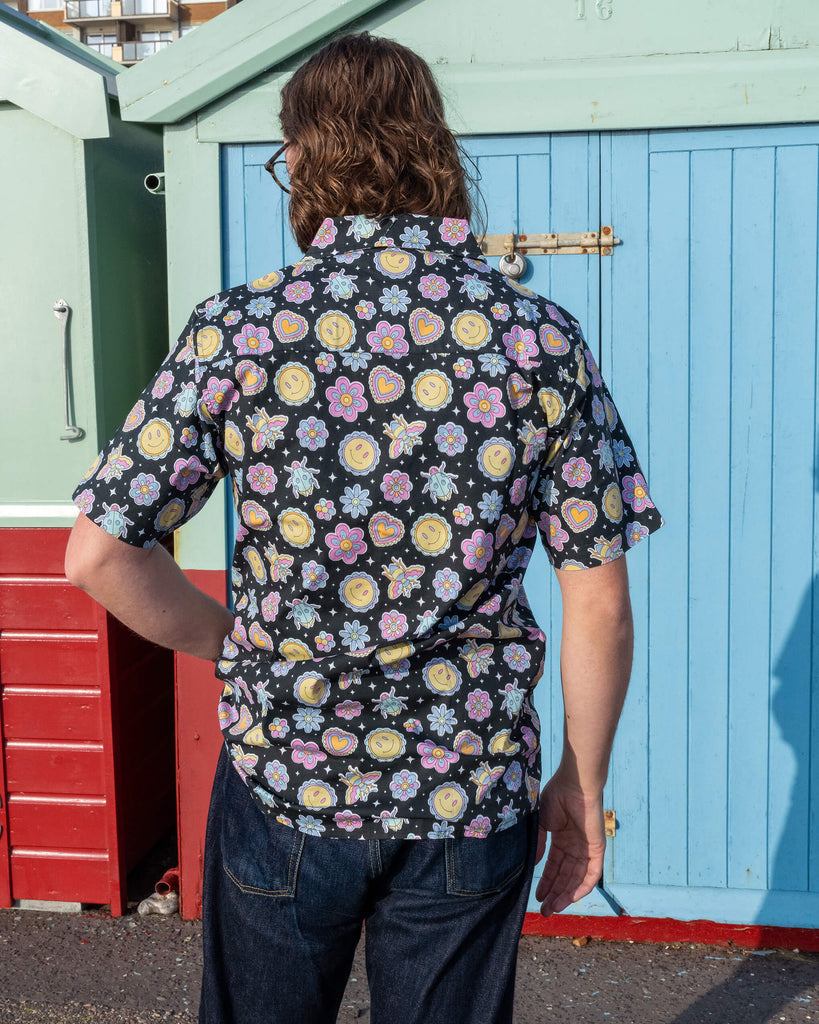 Michael is stood outside in front of a blue beach hut in Hove wearing Run & Fly Bee Happy short sleeve shirt with dark blue jeans. The black shirt has an all over colourful bee, smiley face and flower print. Michael is stood with his back to the camera.