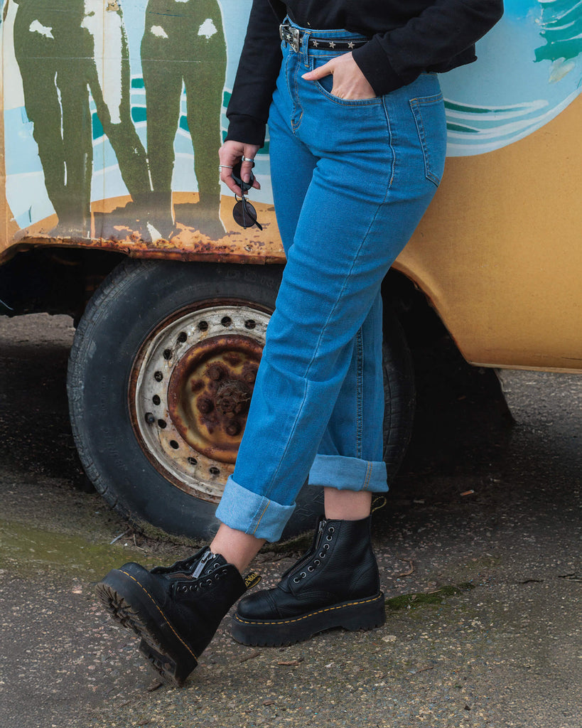 Model Hannah leans against a vintage van, wearing Run & Fly’s blue straight-leg jeans with a rolled-up cuff. She pairs the jeans with chunky black lace-up boots, a black belt with star details, and holds a pair of sunglasses, creating a casual and edgy look.