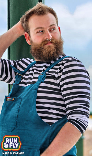Bearded model with silver septum ring wearing a black and white horizontally striped long sleeve t-shirt with ink blue dungarees. They are facing the left and looking over their right shoulder. Photo is cropped from the waist up.