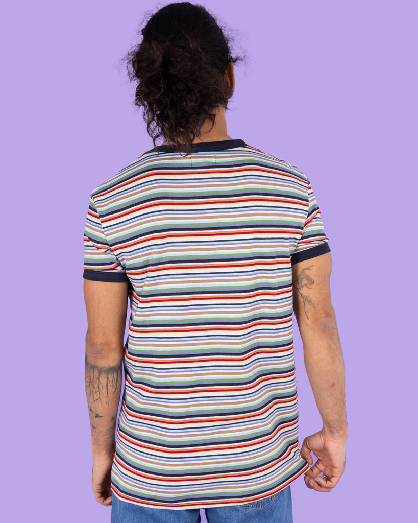 Richard, a hispanic male model with dark hair in a bun and a beard, is stood in a photography studio in Hove in front of a lilac backdrop wearing Retro Multi Striped Ringer Tee with blue jeans. The short sleeved t shirt has horizontal stripes going across it in navy blue, white, orange, brown, yellow, pastel blue and green. Richard is posing with his back to the camera and arms down by his sides.