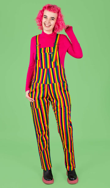 A pink haired model smiling wearing rainbow striped dungarees and a pink top underneath against a green background