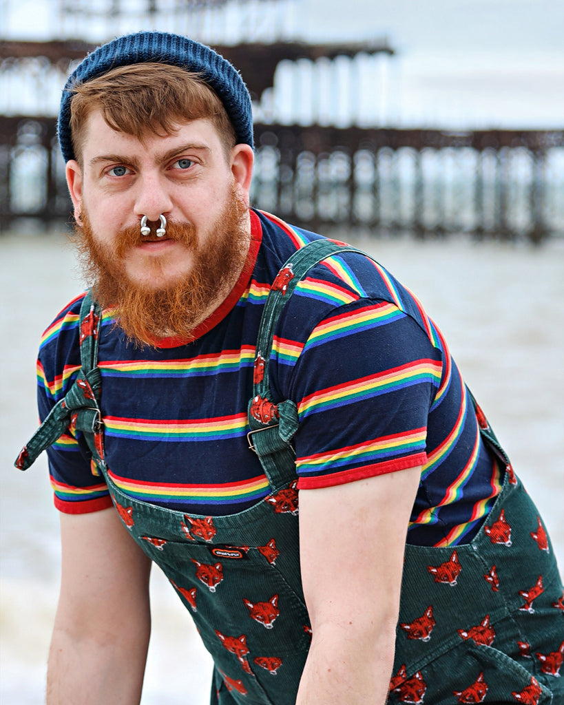 Model standing by the sea wearing a beanie, green fox dungarees and the Retro Rainbow Block Striped T Shirt.