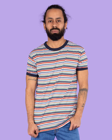 Richard, a hispanic male model with dark hair in a bun and a beard, is stood in a photography studio in Hove in front of a lilac backdrop wearing Retro Multi Striped Ringer Tee with blue jeans. The short sleeved t shirt has horizontal stripes going across it in navy blue, white, orange, brown, yellow, pastel blue and green. Richard is posing facing the camera and is pulling down the bottom of the t shirt. The photo is cropped at the thighs.