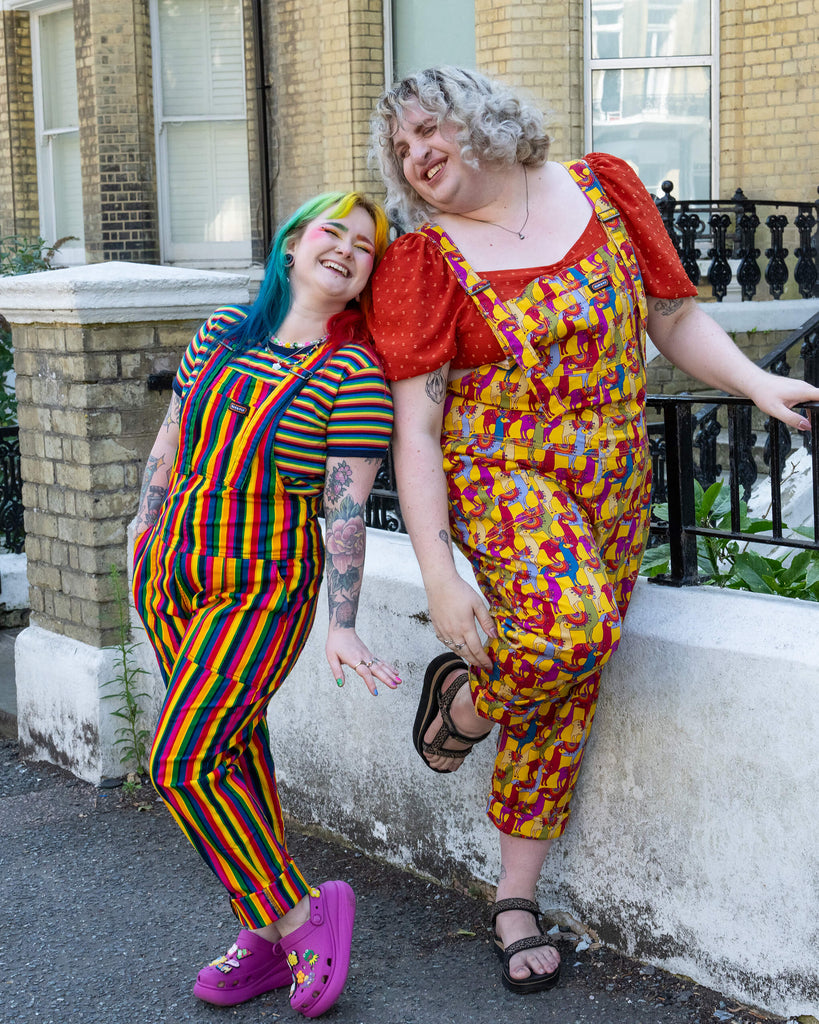 Two people smiling and leaning against a white wall in a cheerful street setting. One wears rainbow-striped dungarees paired with a matching striped shirt and magenta Crocs. The other wears yellow dungarees with a playful llama print and a red puff-sleeved top. They both exude a relaxed and joyful vibe.