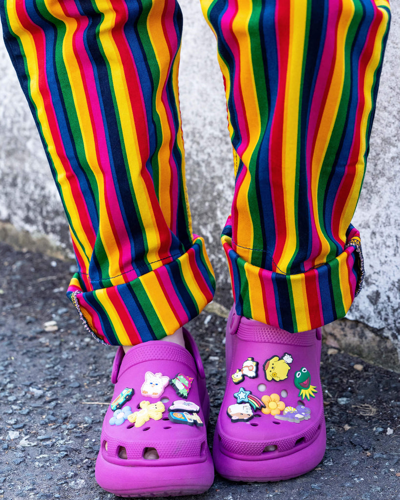 Close-up of bright rainbow-striped dungarees paired with magenta Crocs adorned with fun decorative charms, such as a unicorn, rainbow, and frog. The cuffs of the dungarees are rolled up, showcasing the vibrant vertical stripes in bold colours like yellow, red, blue, green, and purple.