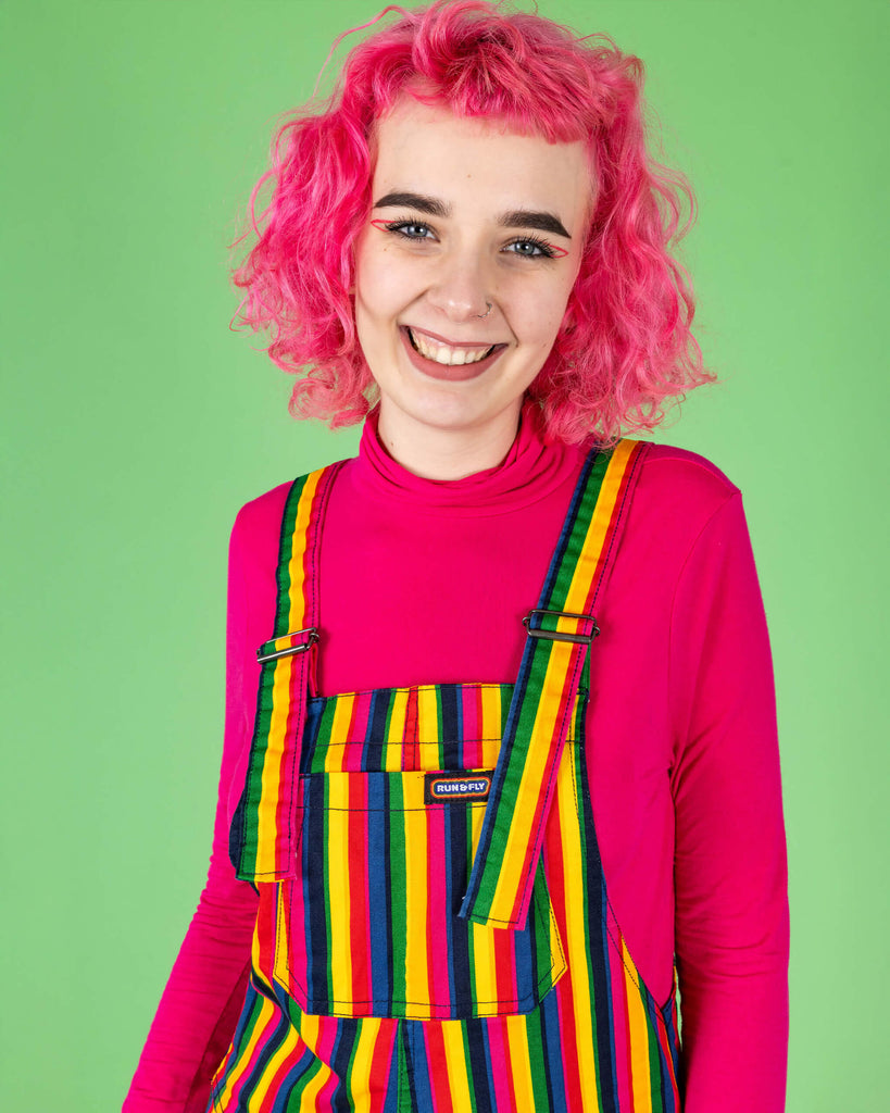 A close-up portrait of a model with pink curly hair wearing rainbow stripe dungarees by Run & Fly. The colourful vertical stripes pop against the bright pink top, with the model smiling brightly against the green background. Run & Fly is a small, ethical, slow fashion company.