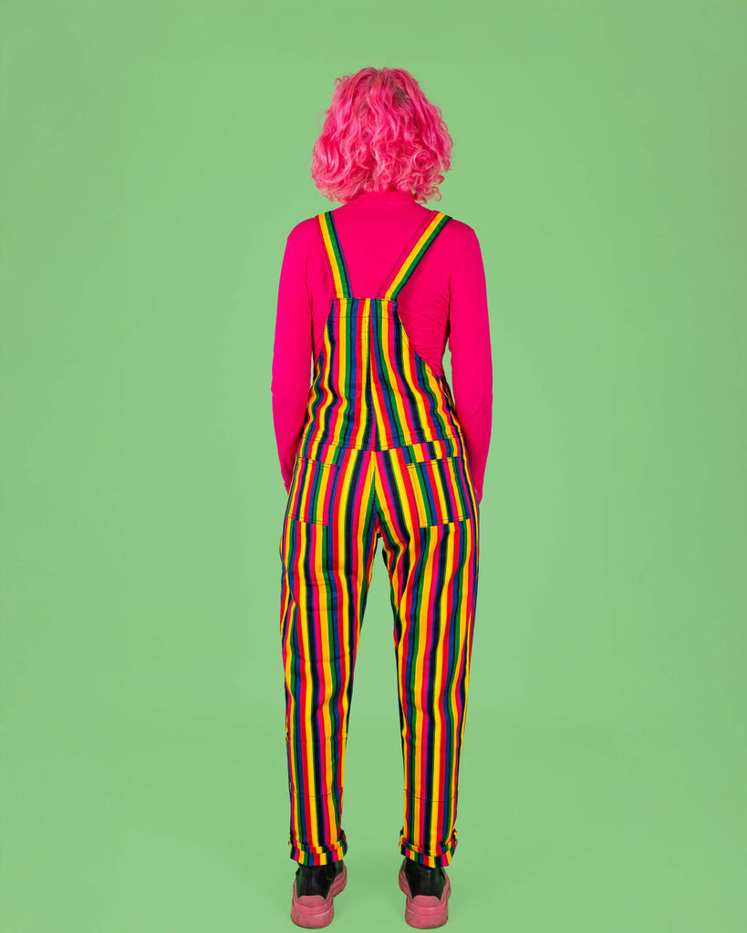 A model with pink curly hair is seen from the back, wearing rainbow stripe dungarees paired with a bright pink top. The bold vertical stripes in various colours stand out against the green background, creating a fun, vibrant look. Run & Fly is a small, ethical, slow fashion company.