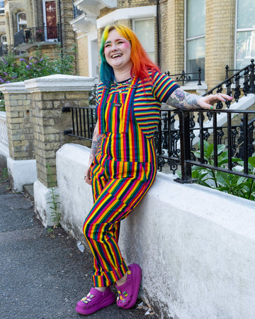 A model with rainbow hair leans against a white wall, wearing matching rainbow stripe dungarees and a T-shirt by Run & Fly. The bright stripes and purple Crocs add a playful vibe to the outfit, with the model smiling and enjoying the outdoor scene. Run & Fly is a small, ethical, slow fashion company.