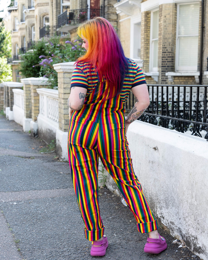 A model with rainbow hair stands outdoors wearing rainbow stripe dungarees by Run & Fly. The dungarees are paired with a matching rainbow-striped T-shirt and purple Crocs. The vibrant outfit creates a colourful, playful look, set against a casual street scene. Run & Fly is a small, ethical, slow fashion company.