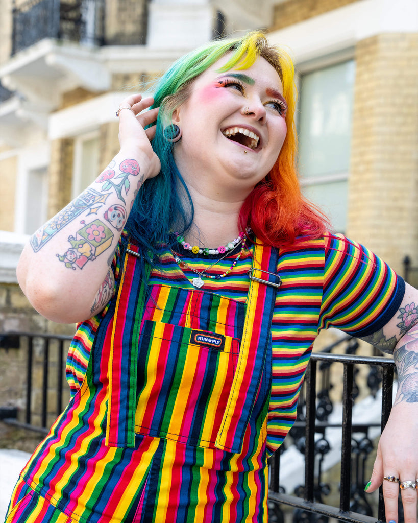 A person laughing and looking off-camera while wearing rainbow-striped dungarees paired with a matching rainbow-striped shirt. Their multi-coloured hair, colourful makeup, and visible tattoos add an extra layer of personality to the bright and fun outfit.