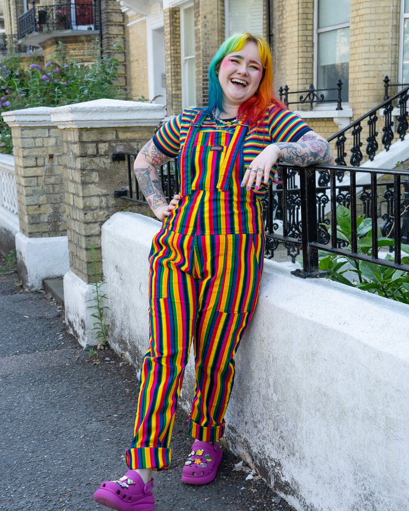 A person standing against a low white wall, wearing rainbow-striped dungarees with a matching rainbow-striped shirt and magenta Crocs. Their multi-coloured hair and playful style complement the vibrant patterns, while they smile brightly in a casual street setting.