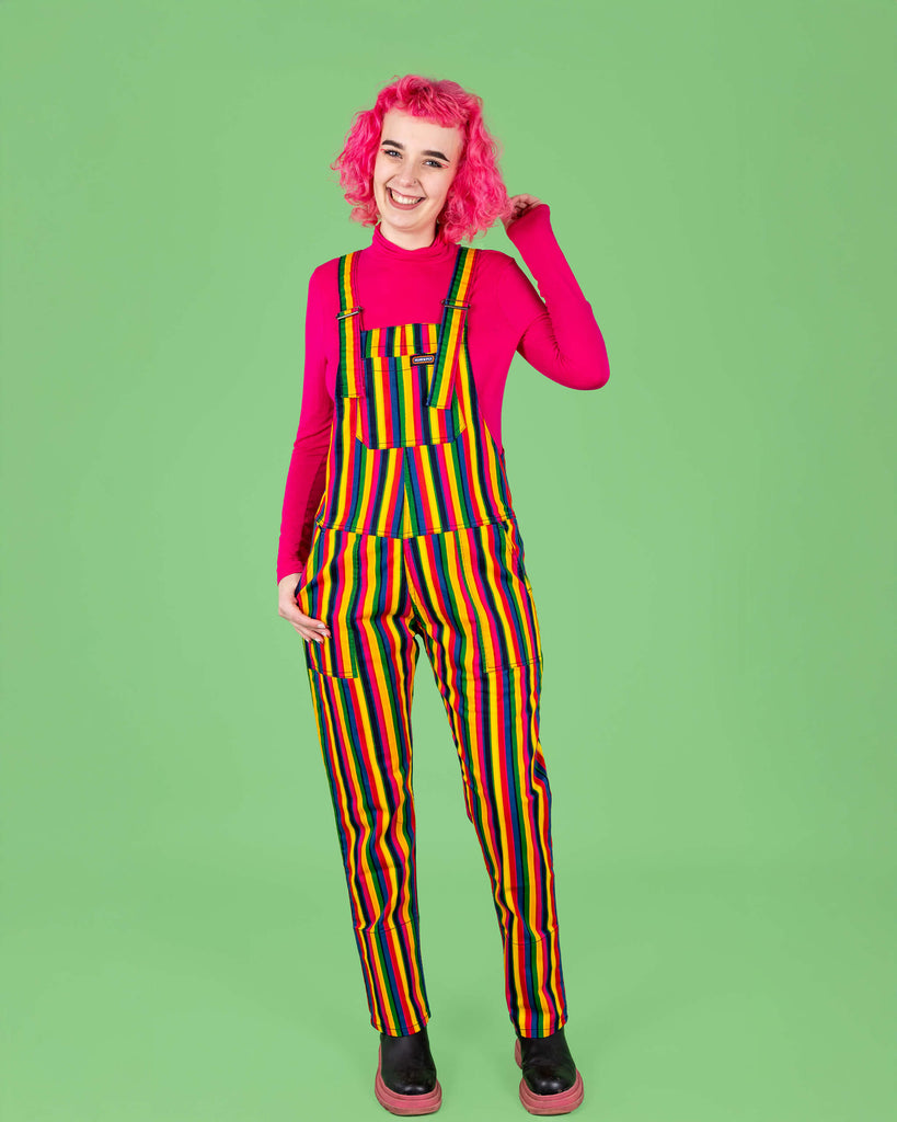 The same model with pink curly hair wears rainbow stripe dungarees by Run & Fly, paired with a bright pink long-sleeve top and black boots. The vibrant rainbow stripes and cheerful expression of the model create a fun, colourful look, set against a bold green background. Run & Fly is a small, ethical, slow fashion company.