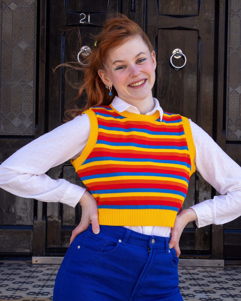 Tasha is a smiley female red haired model with a fringe wearing a white shirt and cobalt jeans with a striped yellow, blue, orange and red tank top. She is stood in a mews in Hove with her hands on her hips