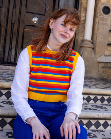 Tasha is a smiley female red haired model with a fringe wearing a white shirt and cobalt jeans with a striped yellow, blue, orange and red tank top from Run & Fly. She is sat on steps outside in Hove with her hair blowing in the wind and hands resting on her knees
